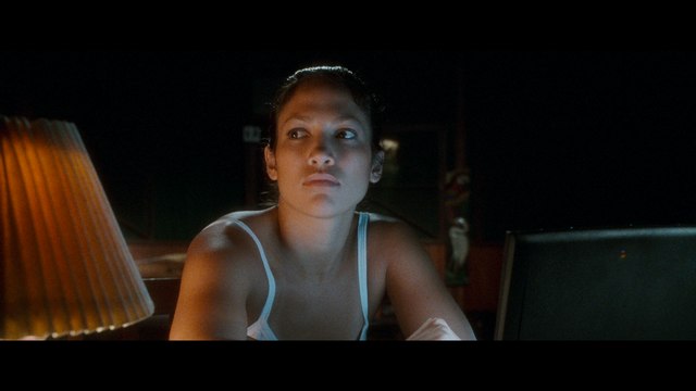 A close-up of a woman sitting at a table, looking intently with a serious expression. A lamp is visible to her left, casting warm light.