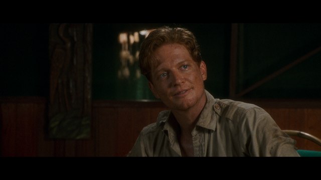 A man with light brown hair and a slight smile, dressed in a button-up shirt, sitting indoors with wooden walls and soft lighting.
