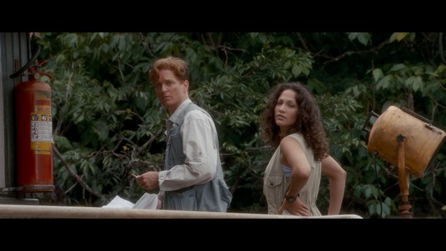 A scene featuring two characters outdoors, surrounded by greenery. One character has red hair and is wearing a light-colored button-up shirt and vest, while the other character has curly hair and is dressed in a sleeveless top paired with overalls.