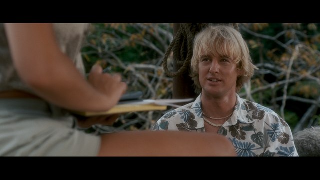 A close-up scene from a movie featuring a man with blonde hair wearing a floral shirt, interacting with a woman sitting nearby. The background includes blurred natural foliage.