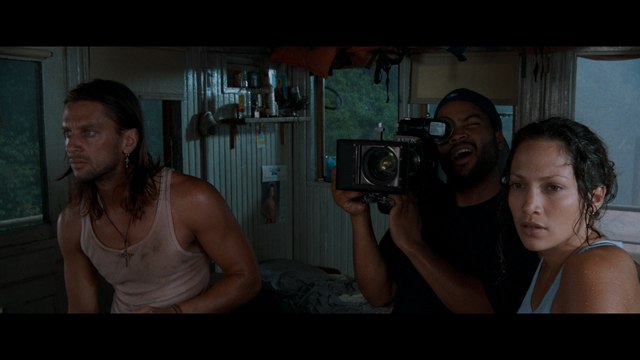 A scene from a film featuring three characters inside a tropical cabin, one holding a camera while the others look on with expressions of concern.