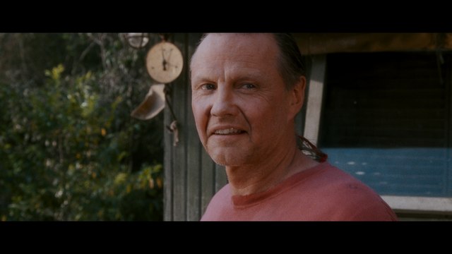 Close-up of a man smiling in front of a rustic cabin, with greenery in the background.