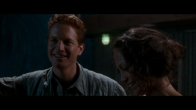 Scene from a film featuring two characters, one male with short blond hair and a gray jacket, smiling at a female character with curly dark hair, both in a dimly lit setting.