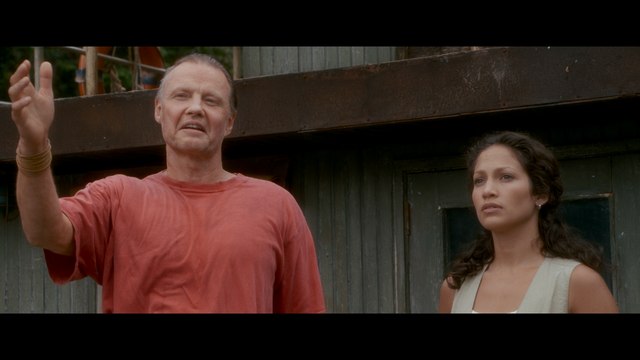 A man in a red shirt gestures while speaking, next to a woman in a light-colored top, both standing in front of a rustic structure.