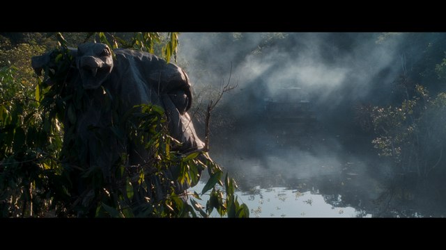 A partially obscured statue of a large creature surrounded by lush greenery and mist above a still body of water.