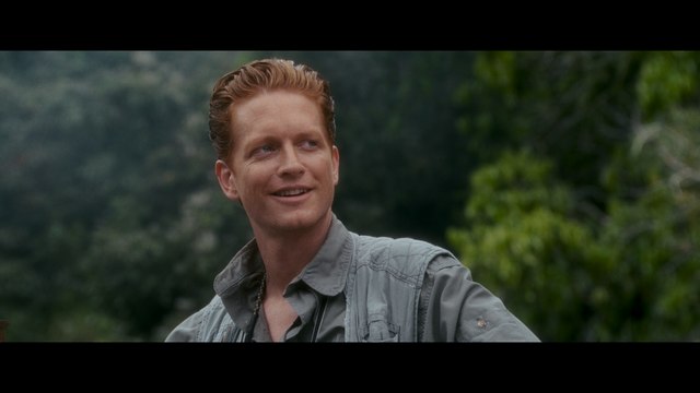 A smiling man with reddish hair wearing a gray shirt, set against a backdrop of lush greenery.