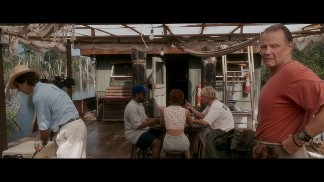 A scene from a boat cabin with several people engaged in conversation, featuring a man in a hat, a woman in a white top, and a man looking intently towards the camera.