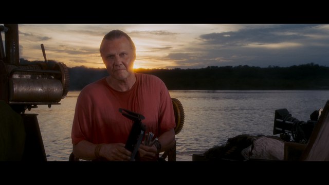 A man in a red shirt holding equipment, standing on a boat near the water during sunset, with a mountainous landscape in the background.