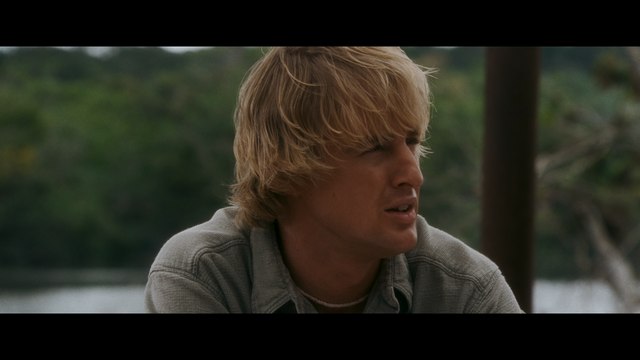 A young man with wavy blonde hair sitting outdoors, looking contemplative with greenery and water in the background.