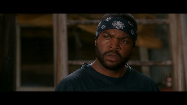 A man with a serious expression wearing a blue bandana and a dark shirt, standing in a rustic setting with wooden structures in the background.