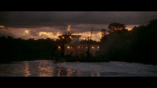 A dark, atmospheric scene featuring a barge on a river during sunset, with dramatic clouds in the sky and lights glowing from the structure.