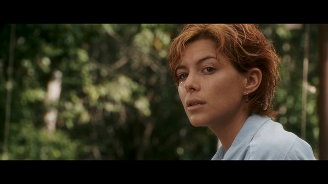 Close-up shot of a woman with short hair, looking contemplative, set against a blurred green background.