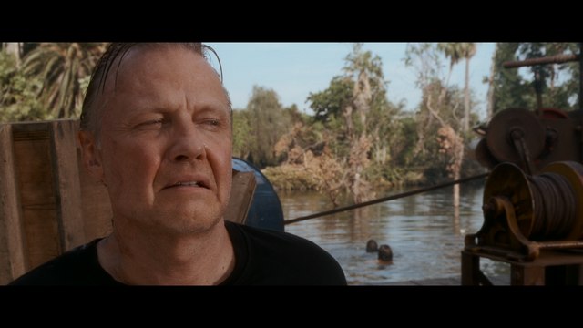 A close-up of a man with an uneasy expression, standing near a river with lush greenery in the background. Two figures can be seen swimming in the water behind him.