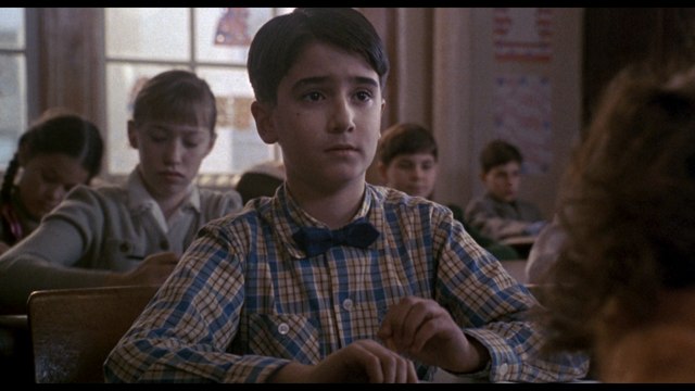 A young boy with dark hair and a checkered shirt sits at a classroom desk, looking serious and contemplative, while other students are visible in the background.