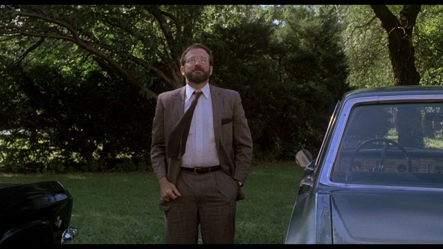 A man in a suit stands in a grassy area near a parked car, surrounded by trees.