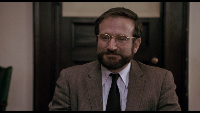 A man sitting at a table wearing a brown suit and glasses, with a slight smile on his face.
