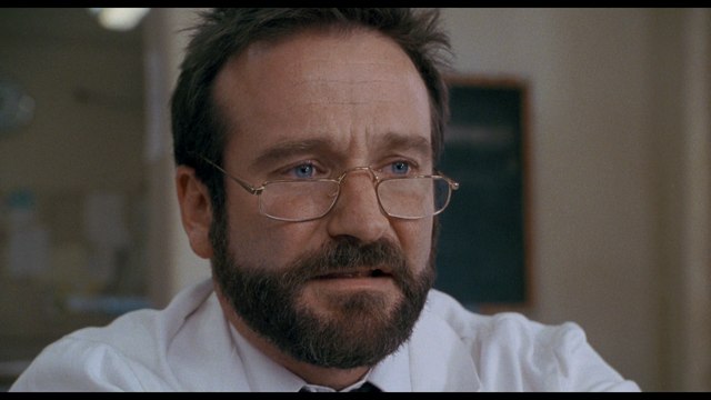 A close-up of a man with glasses and a beard, looking serious or contemplative, in a clinical or office setting.