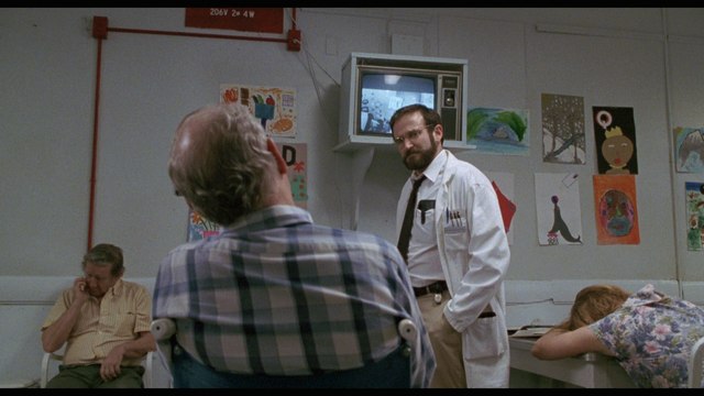 A scene from a movie showing a doctor in a white coat speaking to a patient in a waiting room with artwork on the walls. Other patients are present, one is seated and another is resting their head on a table.