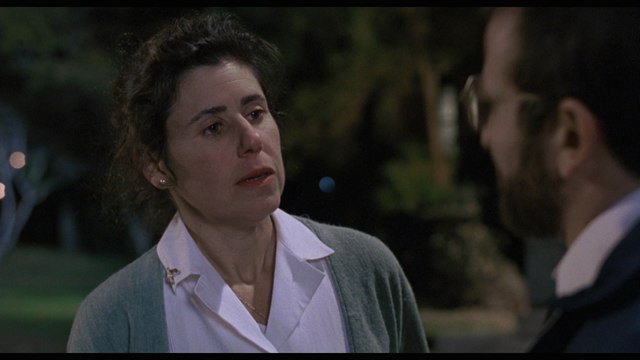 A close-up scene featuring a woman in a white shirt and cardigan, showing a concerned expression while engaging with a man in a dimly lit outdoor setting.