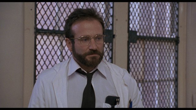 A man with a beard and glasses wearing a white lab coat, standing in a room with a grid-patterned window in the background.