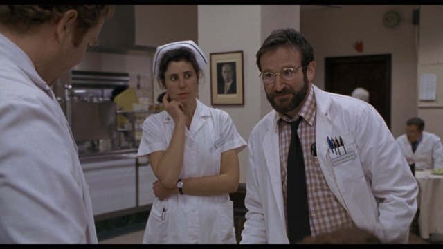 A scene in a hospital kitchen featuring three individuals. A male doctor with glasses and a beard stands to the right, engaged in conversation. A female nurse in a white uniform and cap appears to be thoughtfully listening, while another male staff member is seen in the background.
