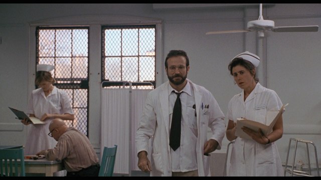 A scene from a hospital featuring a male doctor in a white coat and glasses walking through a corridor, accompanied by two nurses in uniforms. One nurse holds a clipboard while the other stands nearby, and a patient is seated in the background.