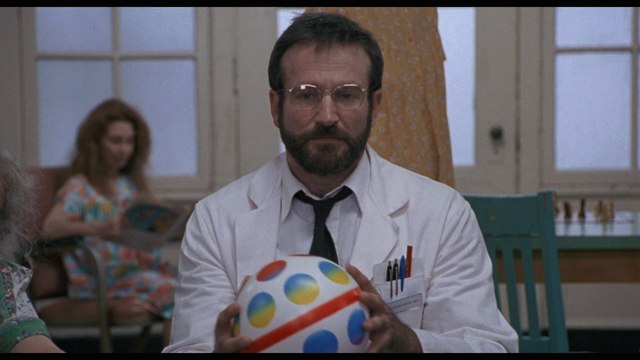 A scene from a movie featuring a man in a white lab coat holding a colorful ball, with a woman in the background seated and reading a magazine.