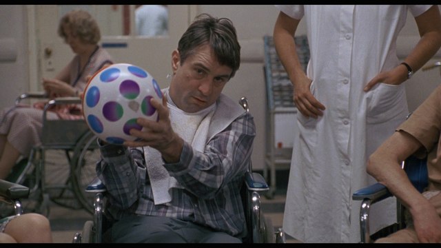 A scene from the movie featuring a man in a wheelchair holding a colorful ball, with a nurse and other patients visible in the background.