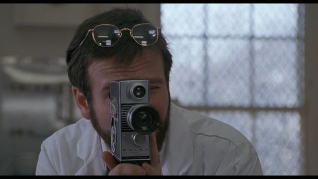 A man wearing glasses holds a vintage video camera, appearing to film in a bright interior space.