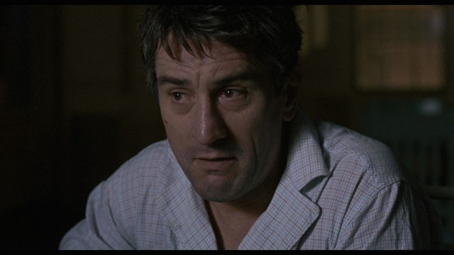 Close-up of a man with short, dark hair and a concerned expression, wearing a light-colored, wrinkled shirt, in a dimly lit room.