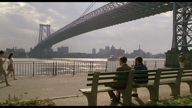 A scene featuring two men sitting on a park bench near a river, with the Williamsburg Bridge in the background and a city skyline visible across the water.