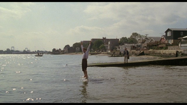 A scene at the water's edge with two individuals. One person stands in shallow water with arms raised, while another figure observes from a wooden structure. Houses and trees are visible in the background under a partly cloudy sky.