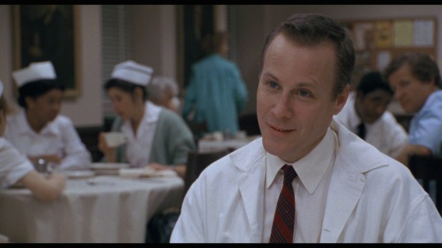 A scene from a movie featuring a man in a white lab coat and tie, smiling while sitting at a table in a cafeteria with several nurses and patients in the background.