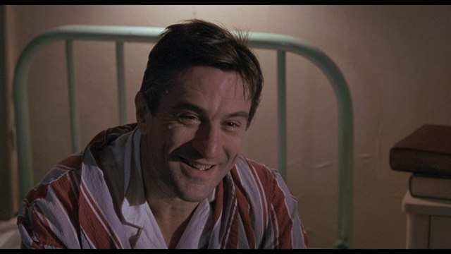 A scene featuring a smiling man wearing a striped pajama top, sitting on a bed with a green metal headboard in the background.