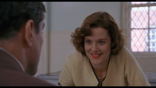 A woman with shoulder-length wavy hair, wearing a light yellow sweater, is smiling and engaging in conversation with a man, portrayed from the side, in a well-lit interior setting.