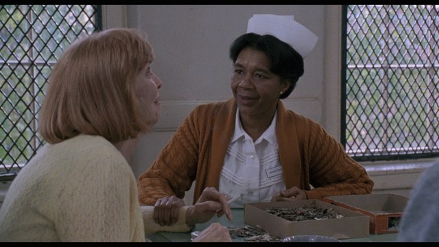 A scene featuring two women sitting at a table. One woman has short hair and wears a nurse's uniform with a white cap, while the other woman has shoulder-length red hair and wears a light-colored sweater. They appear to be engaged in a conversation, with a box of small items on the table between them.