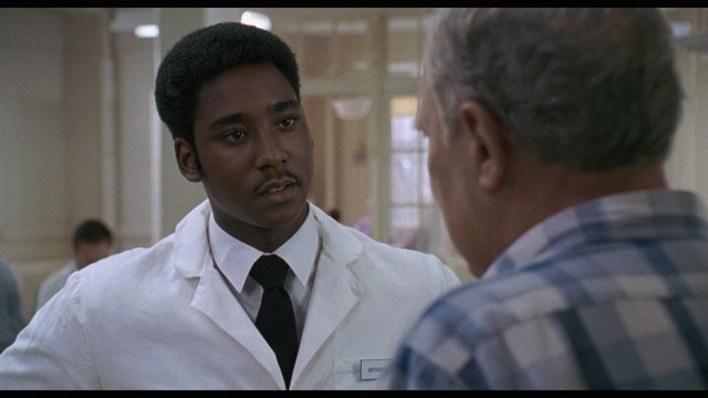 A medical staff member in a white lab coat engages in conversation with an elderly man in a hospital setting.
