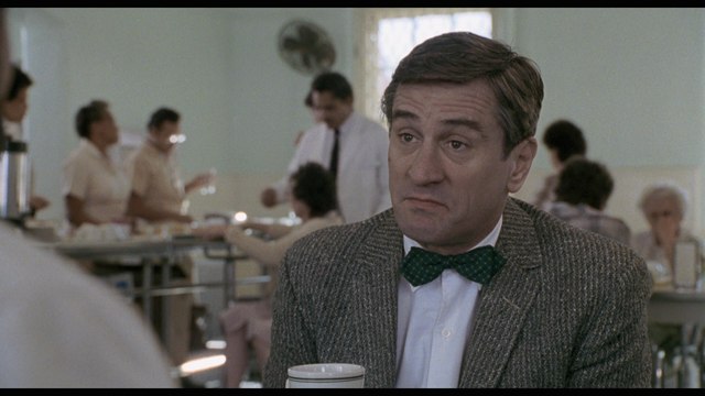 A scene from a film featuring a man in a tweed jacket and bow tie, sitting at a table with a coffee cup, in a cafeteria setting with other people in the background.