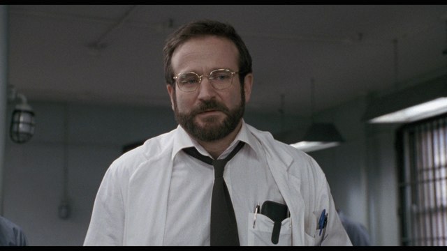 A man with glasses and a beard wearing a white coat and a tie stands in a hospital setting, looking serious.