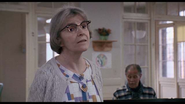 A woman with glasses and gray hair, wearing a patterned shirt and a cardigan, is speaking in a room with a piano and a man in the background.