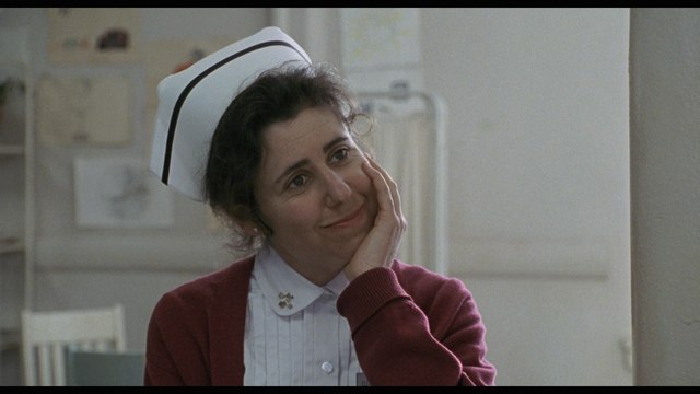 A nurse with a thoughtful expression, wearing a white cap and a red cardigan, in a hospital setting.