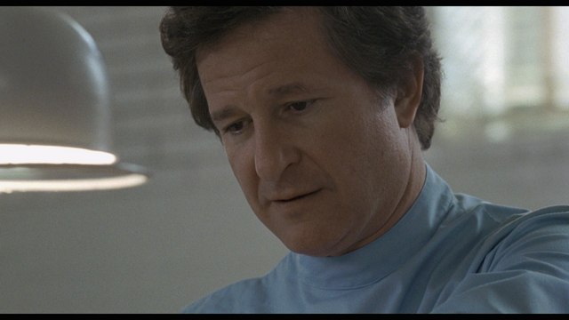 A close-up shot of a man in a blue medical outfit, looking thoughtfully downwards.