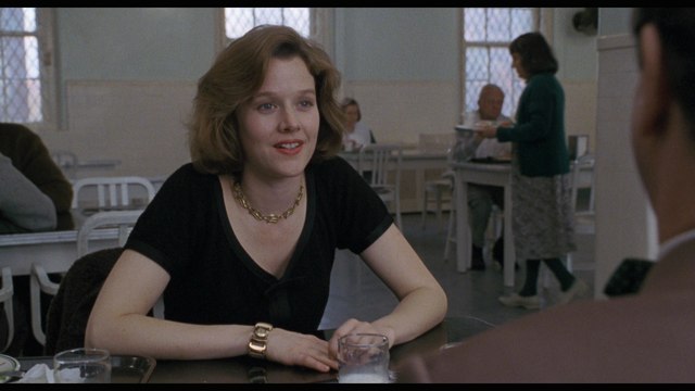 A woman with medium-length brown hair wearing a black top and a necklace sitting at a dining table, smiling while a man sits across from her. The background shows other people in a cafeteria setting.