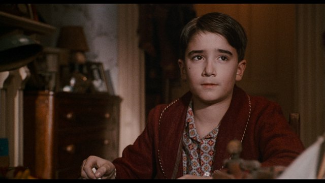 A young boy wearing a red robe and patterned shirt, sitting at a table with a soft expression, in a nostalgic interior setting.