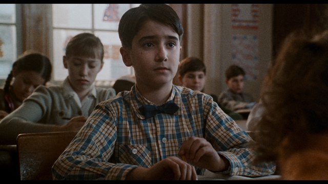 A young boy with a plaid shirt and bow tie sitting in a classroom, looking serious and focused. Other children are seated behind him, engaged in their own activities.