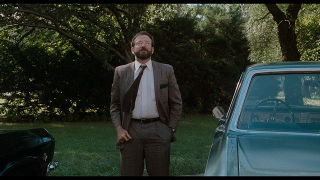A man in a suit stands outdoors, looking up with a thoughtful expression, beside a classic car.