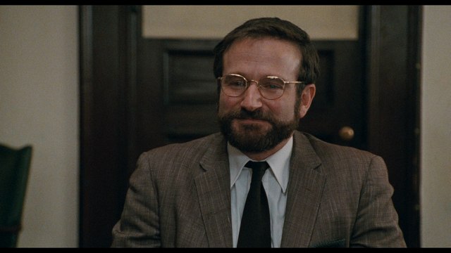 A man in a suit with glasses and a beard is seated at a table, smiling, in a well-lit room with a wooden door in the background.