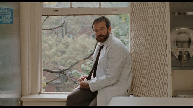 A man in a white lab coat sitting by a window, looking contemplatively outside at trees and buildings.