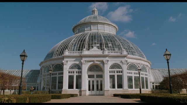 Exterior view of a large glass and white structure, resembling a conservatory or greenhouse, surrounded by neatly trimmed bushes and a clear blue sky.