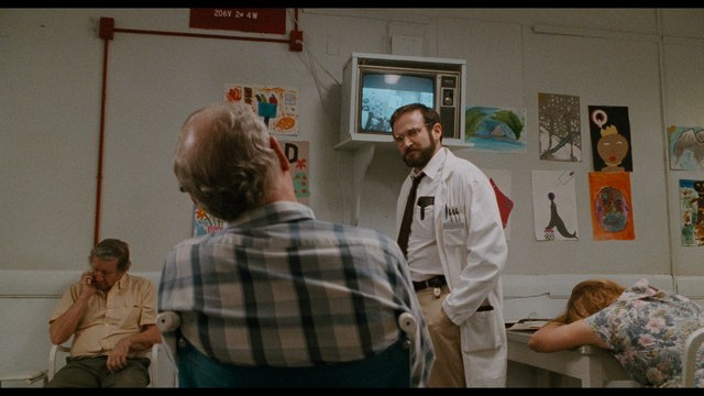 A scene from a medical facility showing a doctor in a white coat talking to a patient seated in a wheelchair, surrounded by other patients and colorful artwork on the walls.